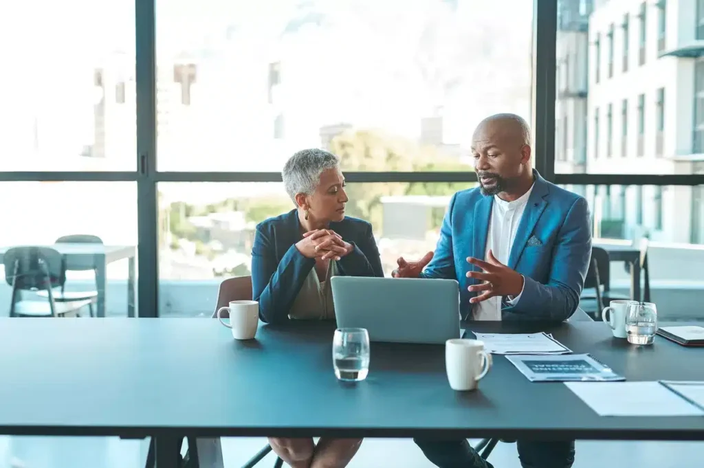 Two professionals sit at a conference table with a laptop, documents, and coffee, having a discussion in a modern office with large windows.