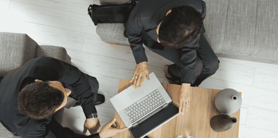 Two people in business attire sit on sofas at a wooden table, looking at a laptop. One person points at the screen. A bag and decorative vases are on the table.