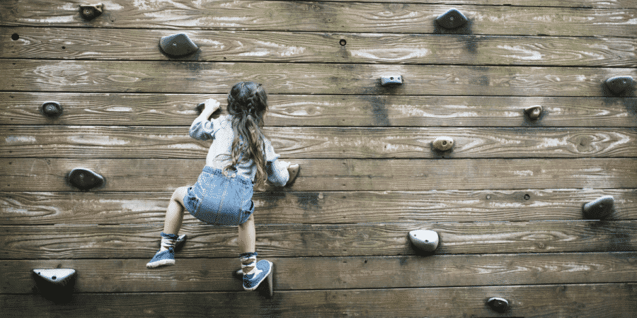 A young girl with long hair climbs an outdoor wooden climbing wall using artificial handholds on a sunny day.