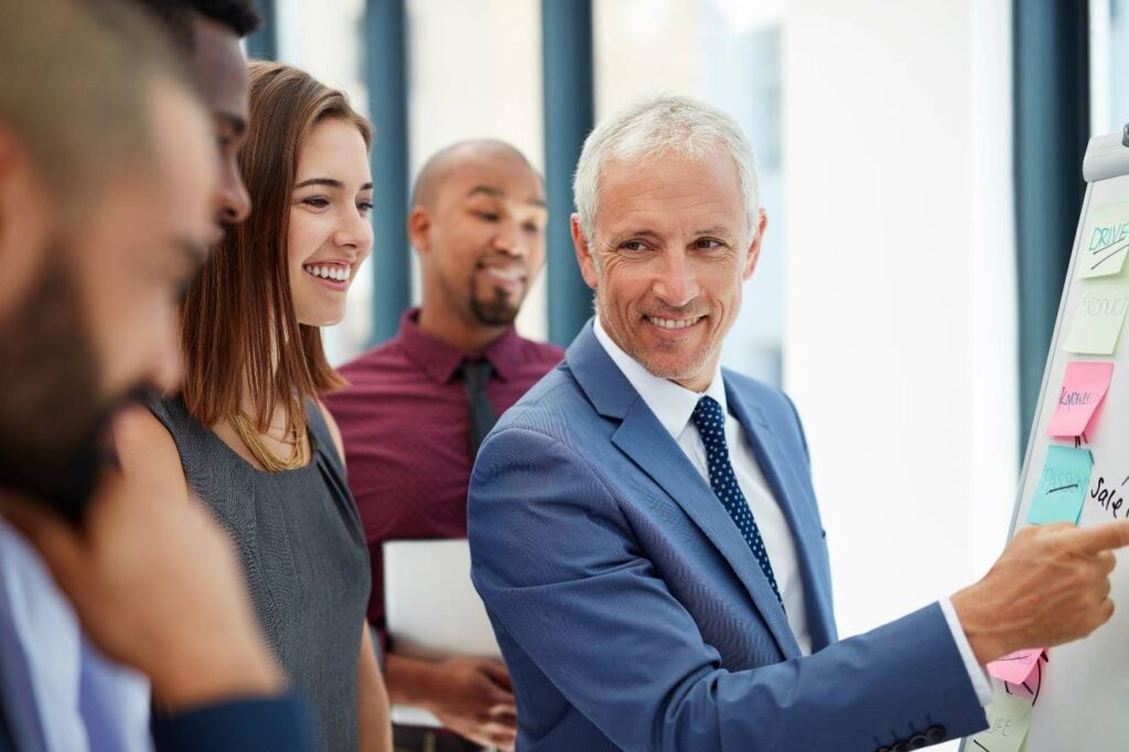 A group of five business professionals, including a man in a blue suit, stand around a flipchart with sticky notes, discussing and smiling.