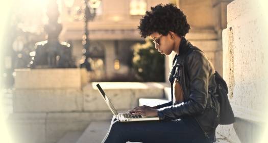 Person sitting on outdoor steps, working on a laptop, wearing glasses and a black leather jacket, with sunlight in the background.