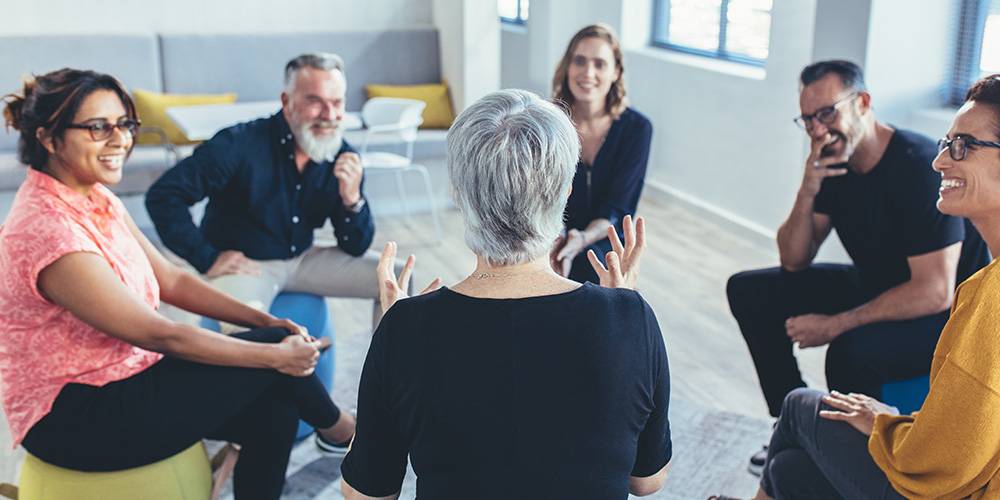 A group of six adults sit in a circle indoors, engaged in a conversation, with one person speaking and others listening and smiling.