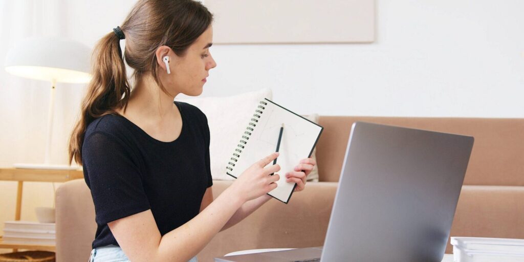A woman with earbuds sits in front of a laptop, holding up a notebook and pen, appearing to explain something during a video call.