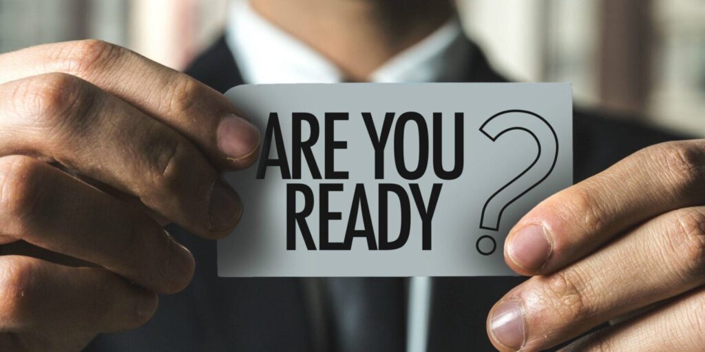 Close-up of a person in a suit holding a card with the text "ARE YOU READY?" in bold letters.