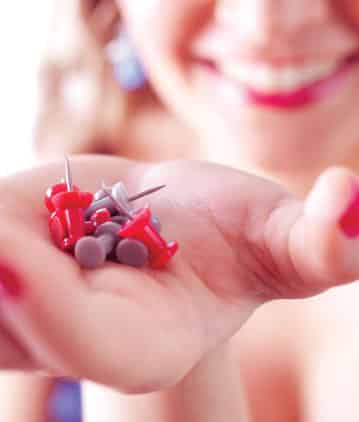 A person holds several red and gray push pins in their open palm, with a blurred smiling face in the background.