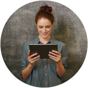 A woman with brown hair in a bun, wearing a blue shirt, stands in front of a gray wall while looking at and holding a tablet.