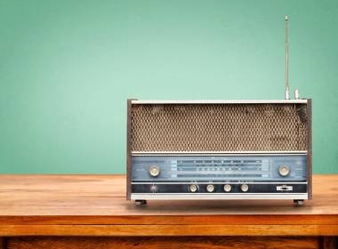 A vintage radio with a metal grille and blue front panel sits on a wooden table against a green background.