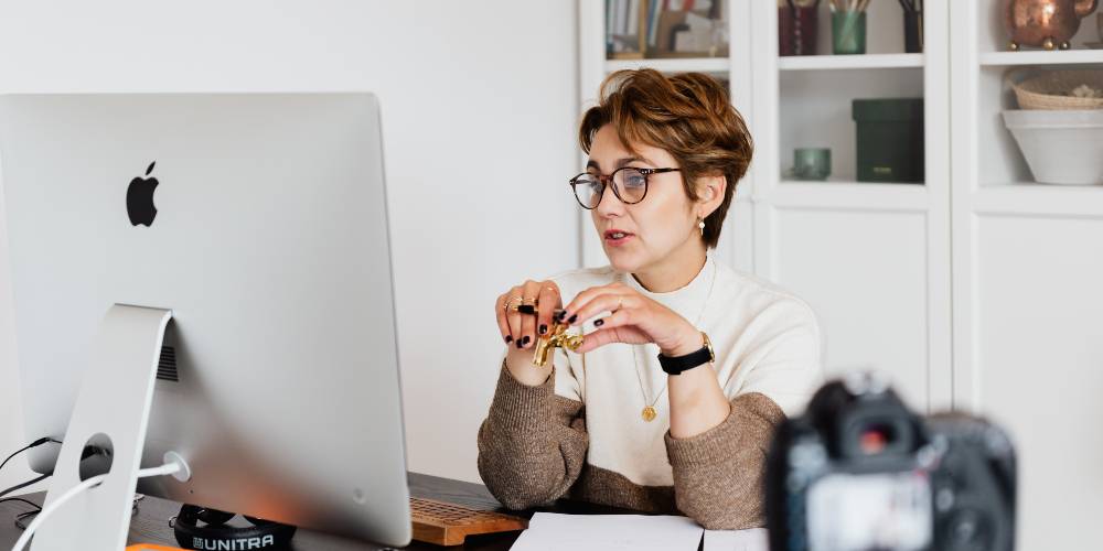 Woman wearing glasses sits at a desk with papers, speaking in front of a large computer monitor and camera in a home office setting.
