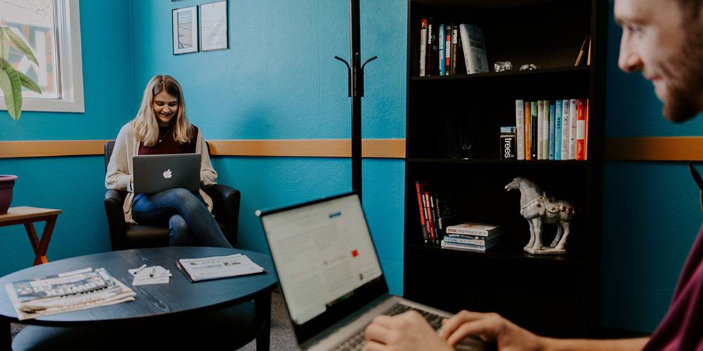 Two people work on laptops in a casual office with teal walls, a bookshelf, and a table with magazines. One person sits at a desk, the other in a chair near the window.