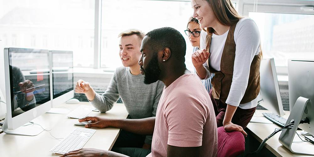 Four people sit and stand around a desk, looking at computer screens and discussing something in an office setting with large windows.