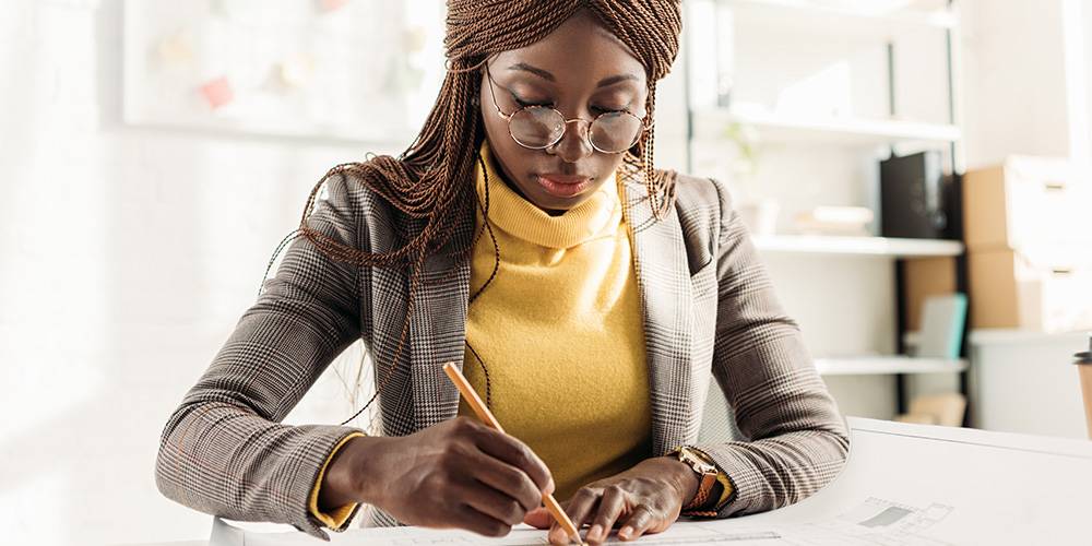 A woman wearing glasses and a yellow turtleneck writes on paper at a desk in a well-lit office.