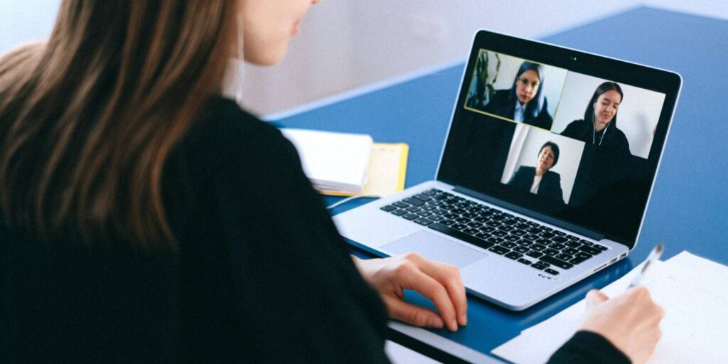 A person takes notes while participating in a video conference with three others displayed on a laptop screen.