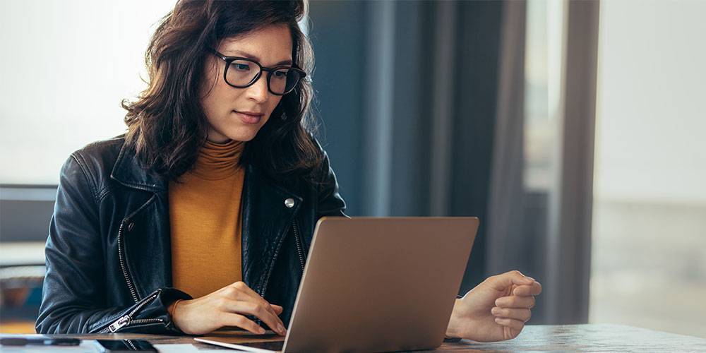 A woman with glasses and dark hair, wearing a leather jacket, sits at a table working on a laptop in a well-lit indoor setting.