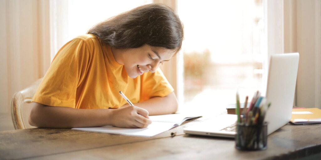 A person in a yellow shirt writes in a notebook at a wooden table, with a laptop and a cup of pens nearby.