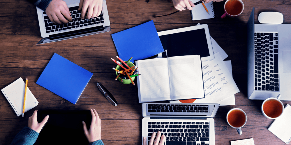 A wooden table with laptops, notebooks, papers, pens, and coffee mugs, with people working and typing, viewed from above.