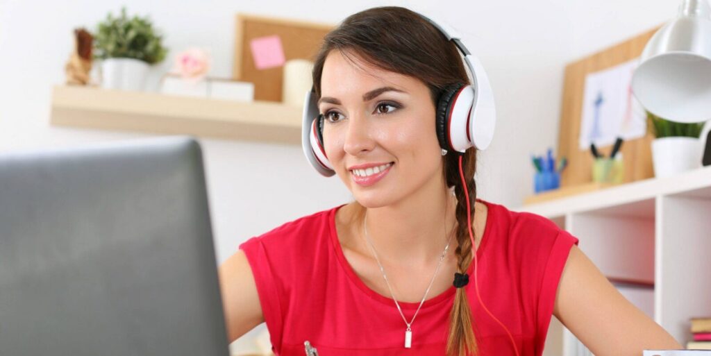 A woman wearing headphones and a red shirt sits at a desk, looking at her laptop and smiling, with office supplies and shelves in the background.