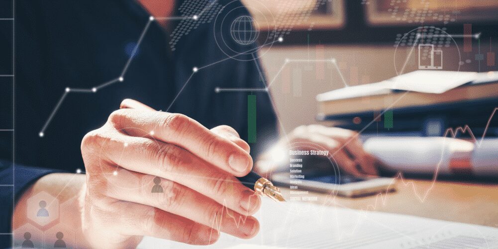 A person holds a pen over paperwork at a desk with digital business and data graphics overlaid, suggesting analysis or strategy planning.