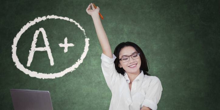 A smiling woman with glasses raises her hand in front of a chalkboard with an "A+" written inside a circle.