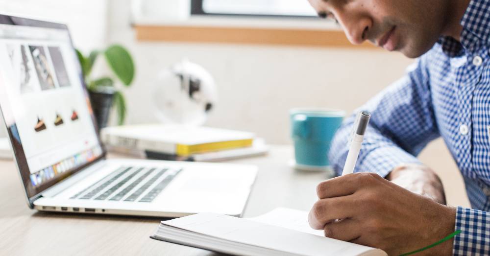 A person writes in a notebook at a desk with an open laptop, books, a blue mug, and a small plant nearby.