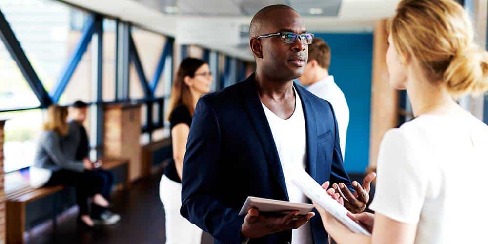 Two people stand indoors having a conversation, while others sit and talk in the background. One person holds a tablet, and another holds documents.