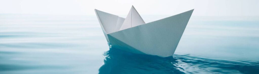 A white paper boat floats on calm, blue water with a light, hazy sky in the background.