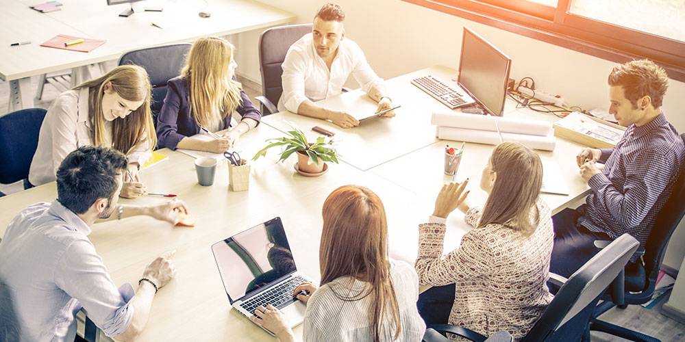 Seven people sit around a table in a bright office, working with laptops, papers, and notebooks, engaged in conversation and collaboration.