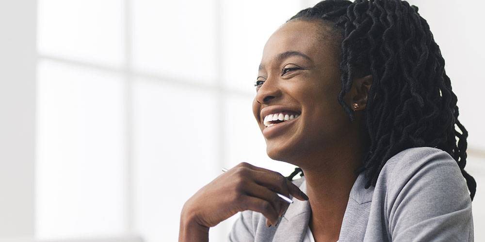 A woman in a light gray blazer smiles while sitting indoors near a large window with natural light.
