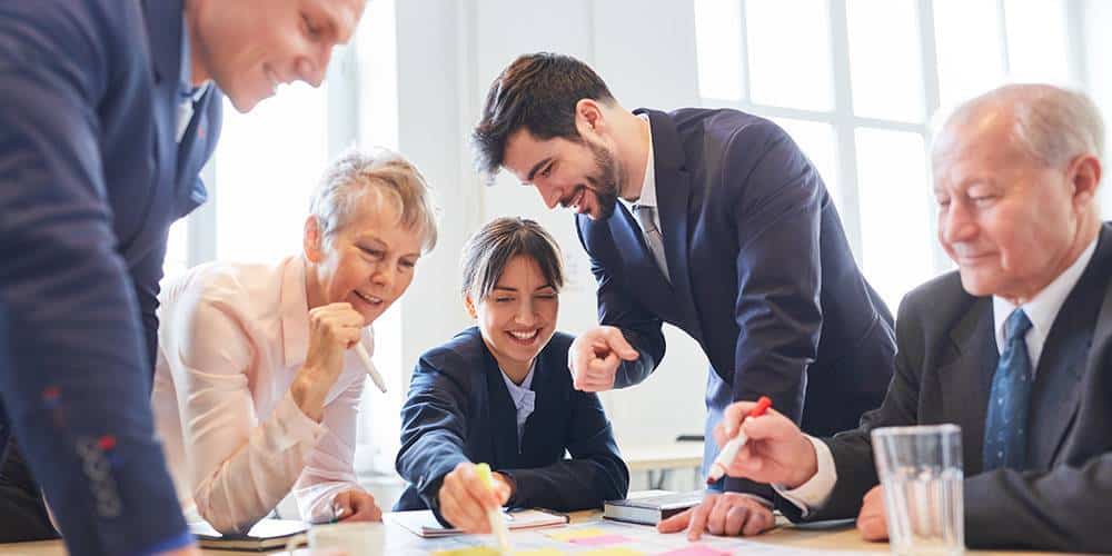 Five people in business attire collaborate around a table, smiling and discussing notes and papers in a bright office setting.