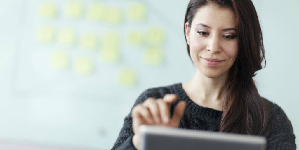 Woman wearing a dark sweater uses a tablet device, with blurred sticky notes on a whiteboard visible in the background.