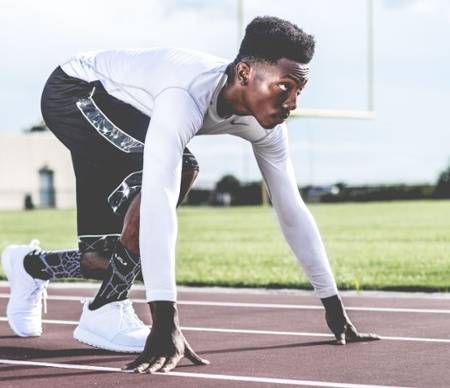 A male athlete in a white shirt and black shorts is in a crouched starting position on a running track, preparing to sprint.