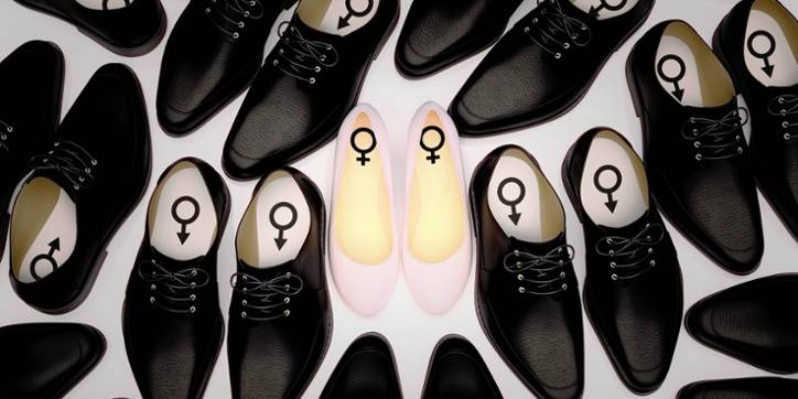 Several pairs of black men’s dress shoes surround a single pair of pale pink women’s flats; male and female symbols are visible inside the respective shoes.