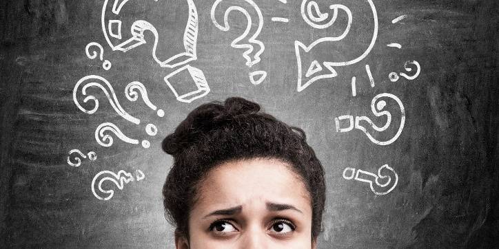 A person looks upward with a confused expression; chalk-drawn question marks surround their head on a blackboard background.