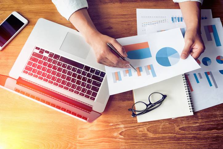 Person analyzing printed charts with graphs at a desk with a laptop, smartphone, eyeglasses, and papers.