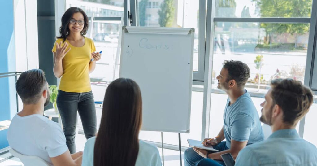 A woman stands by a flip chart presenting to four seated people in a bright, modern office setting.