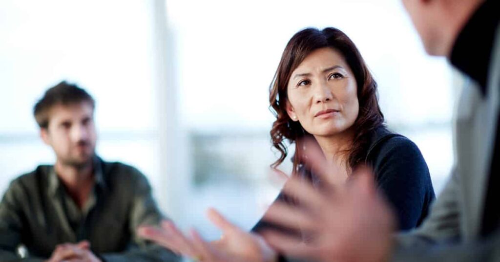 Three people are having a serious discussion at a table; a woman in the center is listening intently to a man speaking in the foreground.