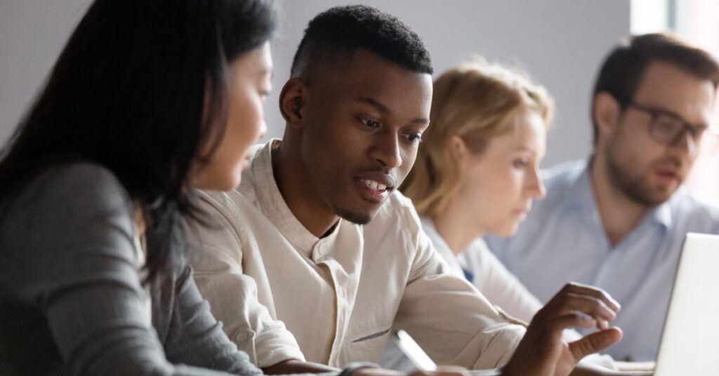 Four people sit at a table working together, with two in the foreground discussing something on a digital device and two more focused on their tasks in the background.