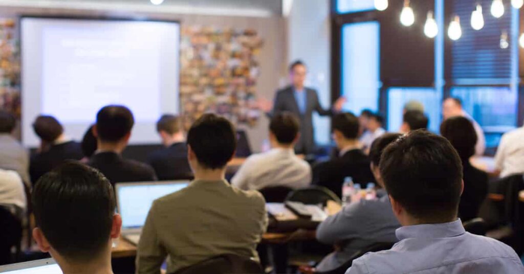 A group of people sit facing a presenter who is speaking at the front of a conference room with a projected screen in the background.