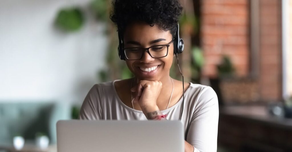A person wearing glasses and headphones smiles while looking at a laptop in a bright indoor setting.
