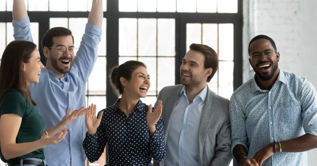 A group of five people stand together indoors, smiling and laughing, with one person raising their arms in celebration.