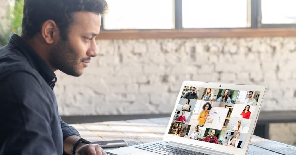 A man sits at a table and looks at a laptop screen displaying a video conference with multiple participants.