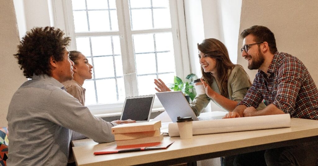 Four people sit around a table with laptops, books, and papers, engaged in a discussion in a bright room with large windows.