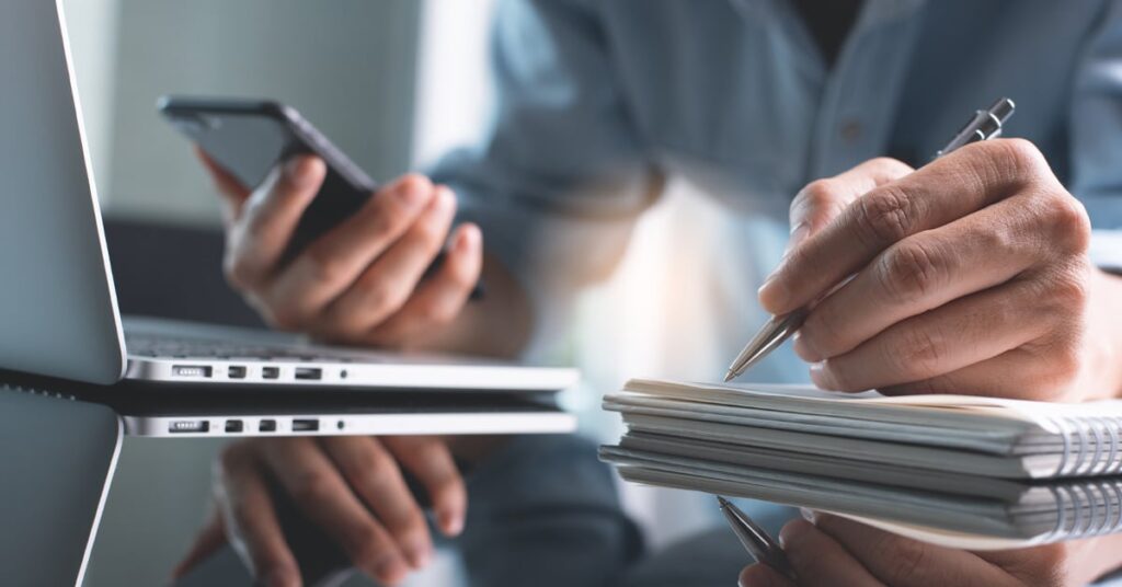 Person working at a desk with a laptop, holding a smartphone in one hand and writing in a notebook with the other hand.