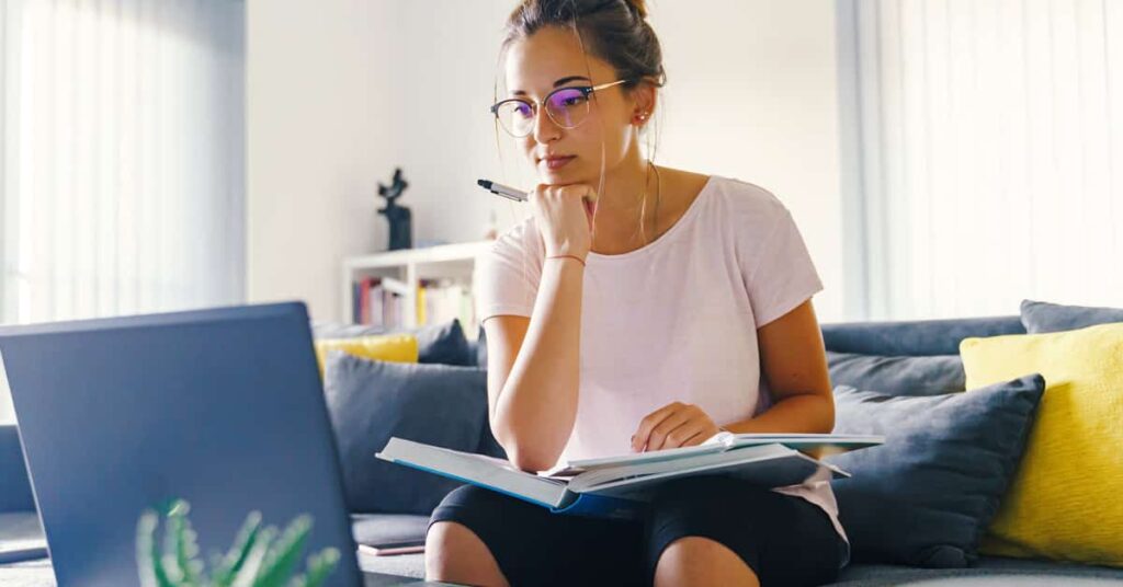 Woman sitting on a couch, holding a pen and open notebook, looking at a laptop screen with a focused expression.