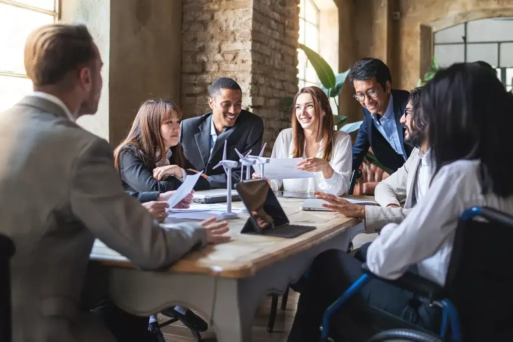 A group of diverse professionals sit around a table in a meeting room, discussing documents and smiling during a collaborative business meeting.