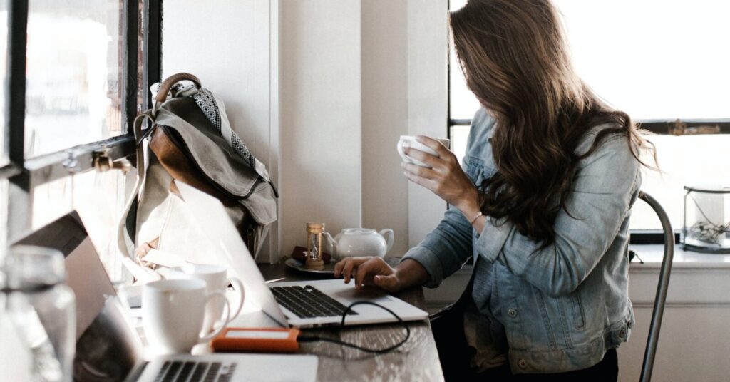 A person with long hair sits at a table working on a laptop while holding a cup, with a backpack and other items nearby.