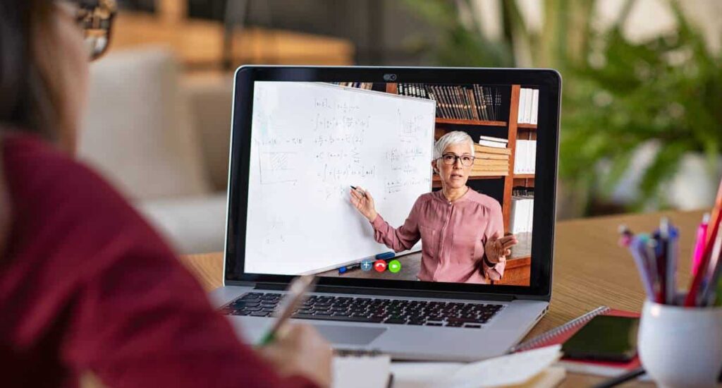 Person attends an online class, watching a teacher explain math concepts on a whiteboard during a video call, with study materials on the desk.