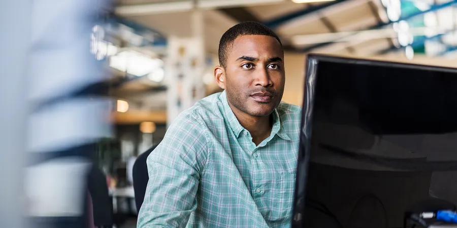 A man in a green plaid shirt sits at a desk, focused on a computer monitor in a modern office setting.