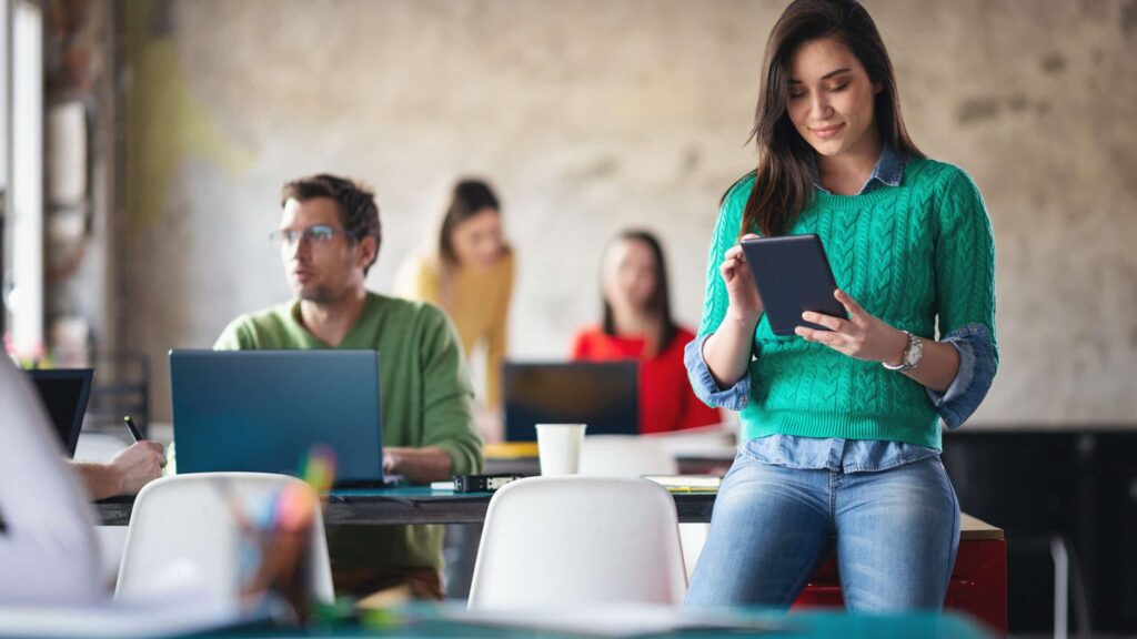 A woman in a green sweater uses a tablet while others work on laptops in a modern, informal office setting.