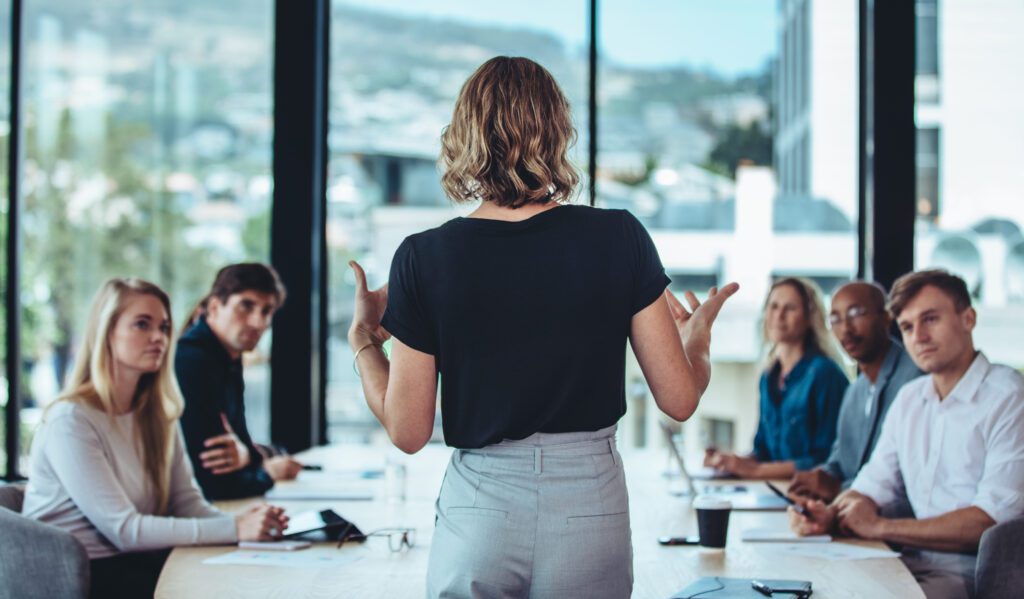 A woman stands addressing a group of five people seated around a conference table in a modern office with large windows.
