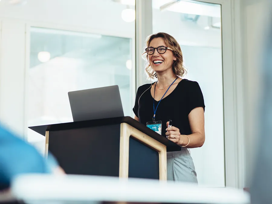 A woman wearing glasses stands at a podium, smiling while giving a presentation with a laptop in front of her.